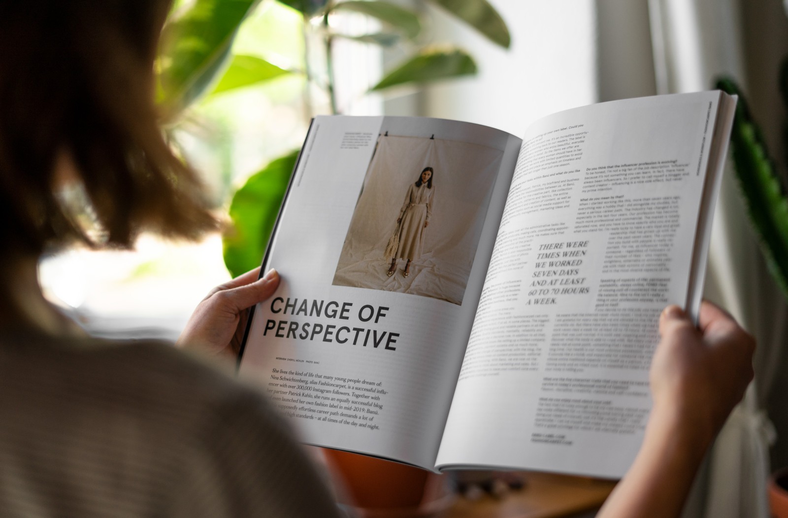 An atmospheric photo of a person reading an open issue of J'N'C magazine in front of a window with houseplants; the visible spread features the cover story “Change of Perspective,” including a fashion portrait and multi-column text
