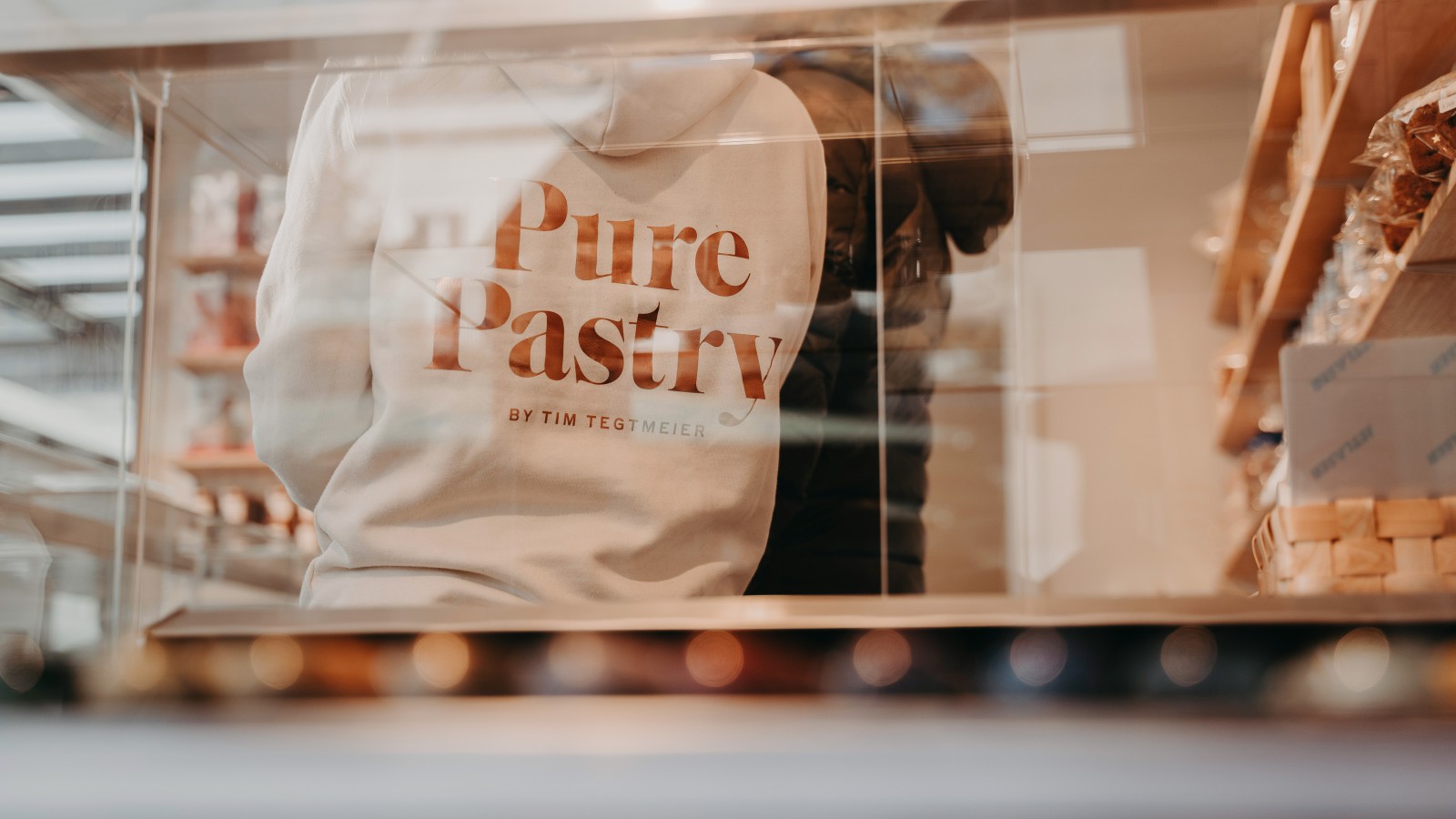 An employee wearing a white Pure Pastry-branded sweater, photographed from outside as they enter the patisserie