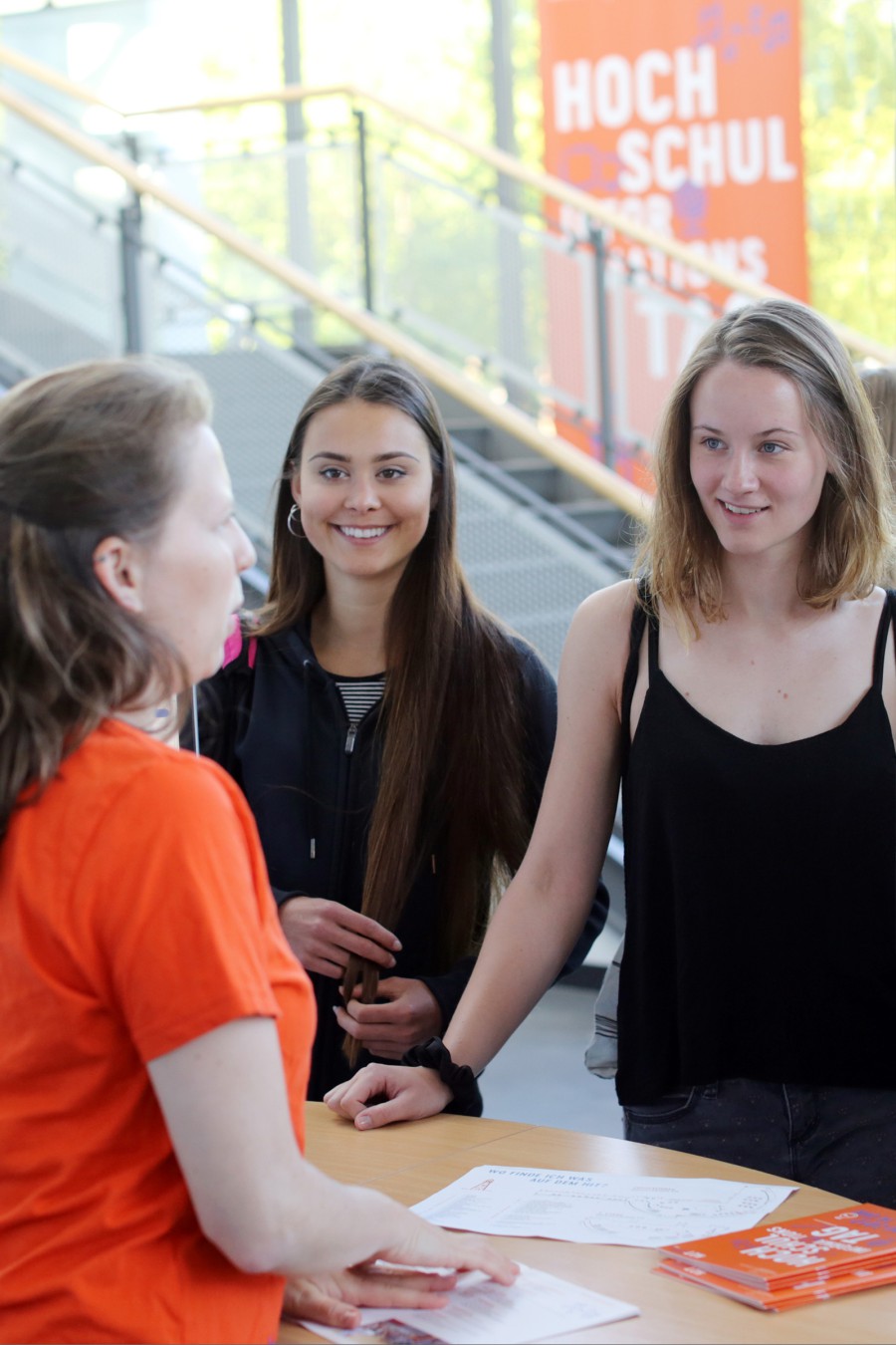 Two high school students are standing at a table talking to a college counselor wearing an orange HIT T-shirt and holding program booklets; in the background is a blue HIT roll-up banner